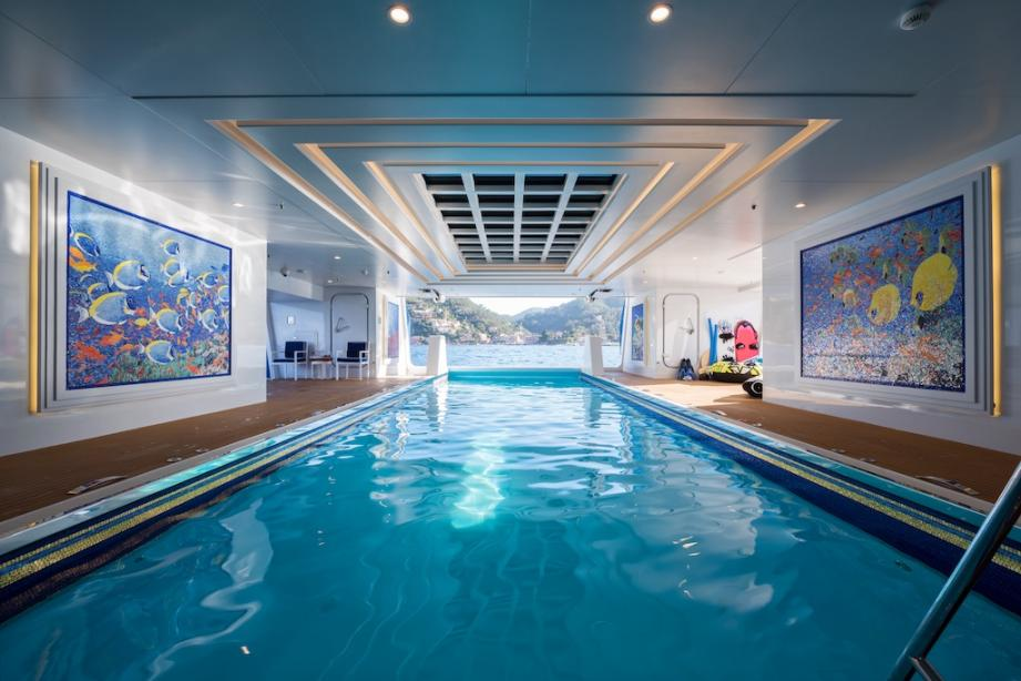 Indoor 25-meter swimming pool on the Dilbar mega yacht with marine-themed artwork and a view of the coastline