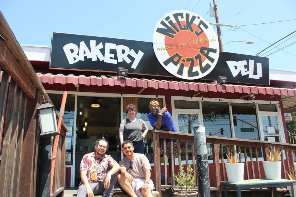 Nick's Pizza Oakland storefront with bakery and deli signage and four people posing outside